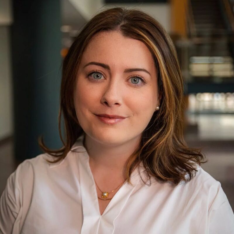 The image shows a woman with fair skin, blue eyes, and brown hair with blonde highlights. She is wearing a white collared shirt and a delicate gold necklace. She is smiling slightly, and the background appears to be an office or public building with blurred architectural details.